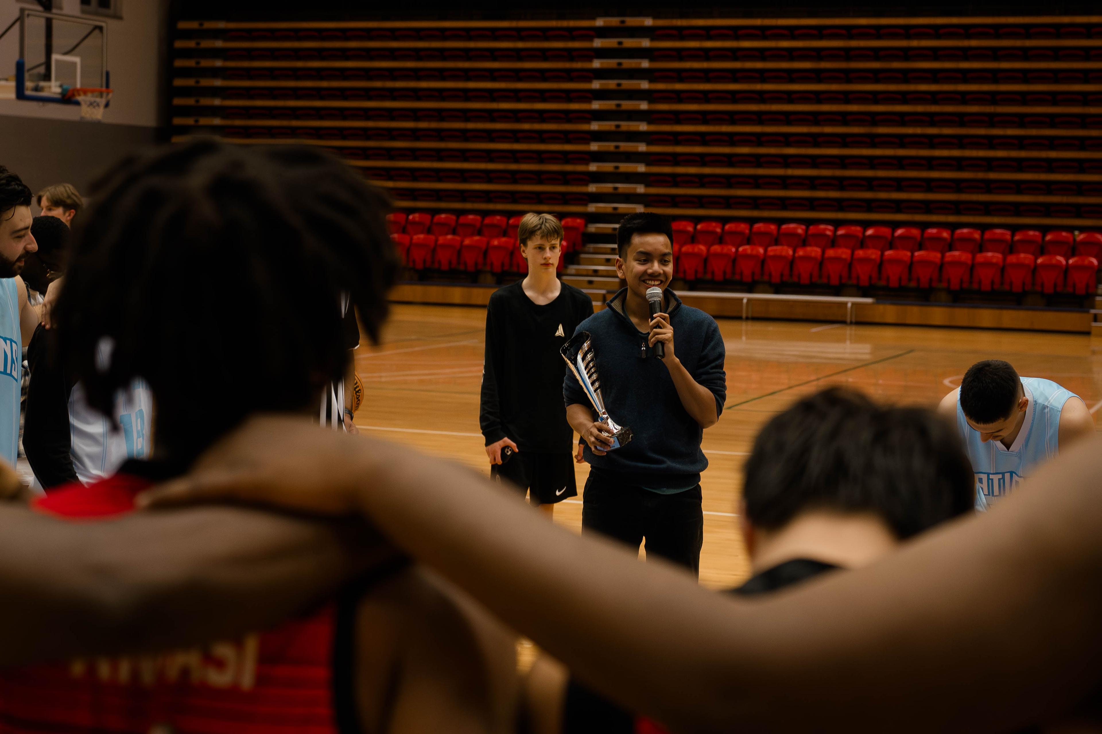 Man holding a trophy and speaking into a microphone as players gather in a huddle.