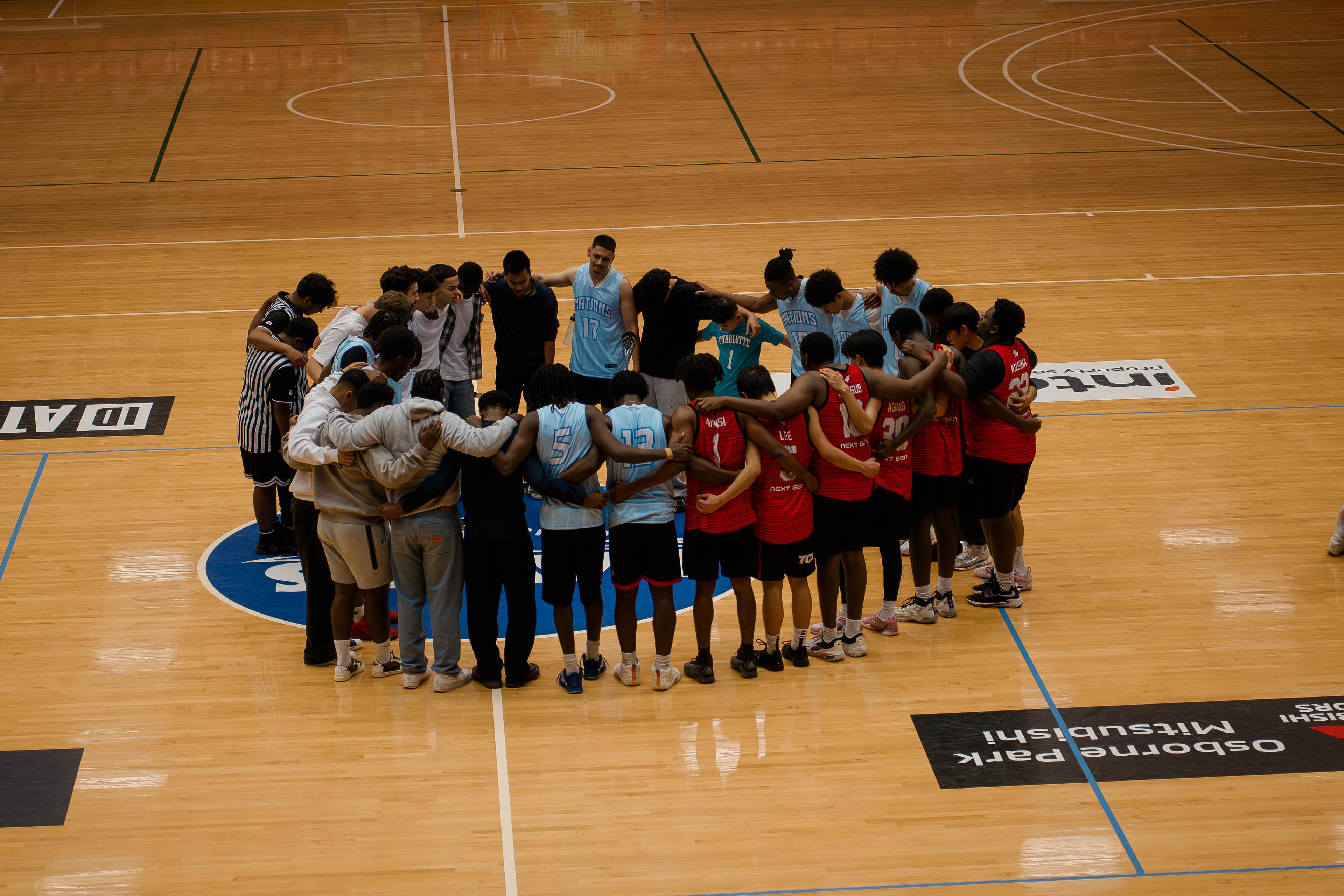Two teams and officials gather in a large circle at centre court after a game.