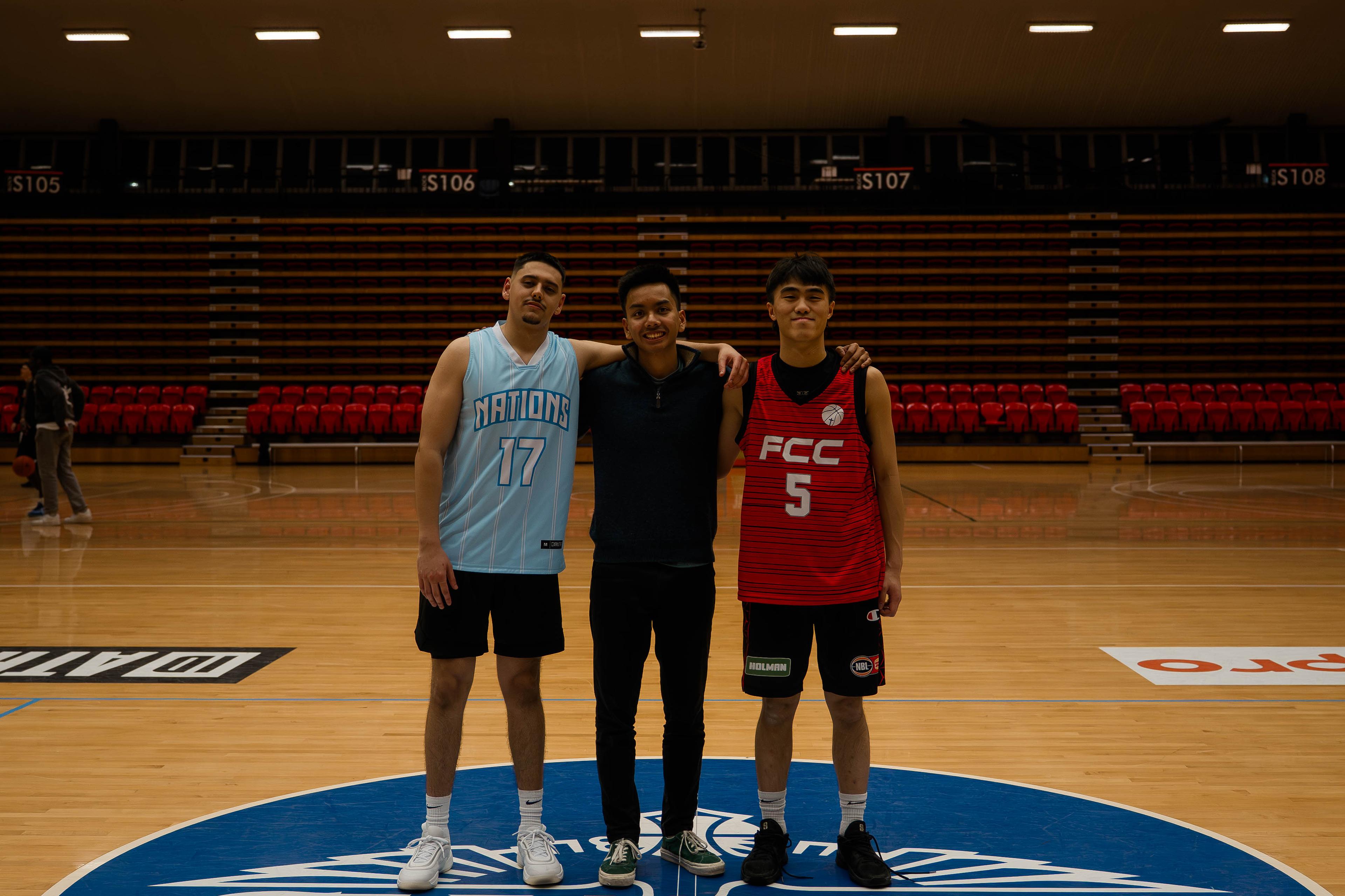 Three people pose arm in arm at centre court, two wearing team jerseys.